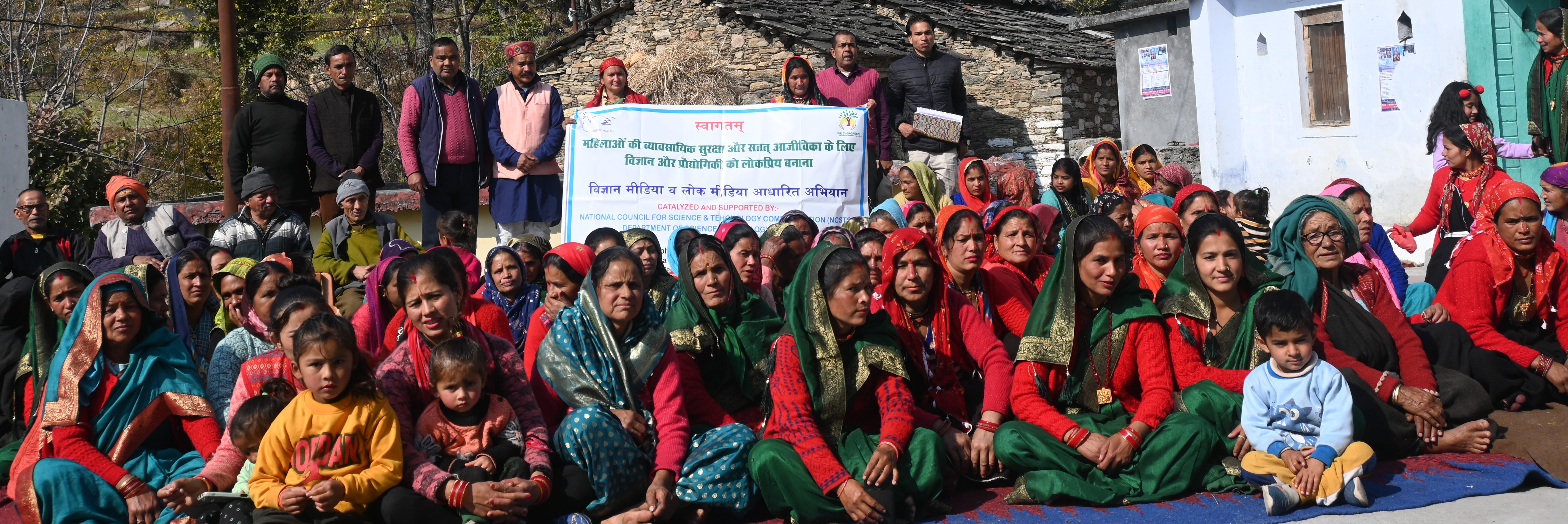 Women and organizers gather outdoors behind a community programme banner at Simli.